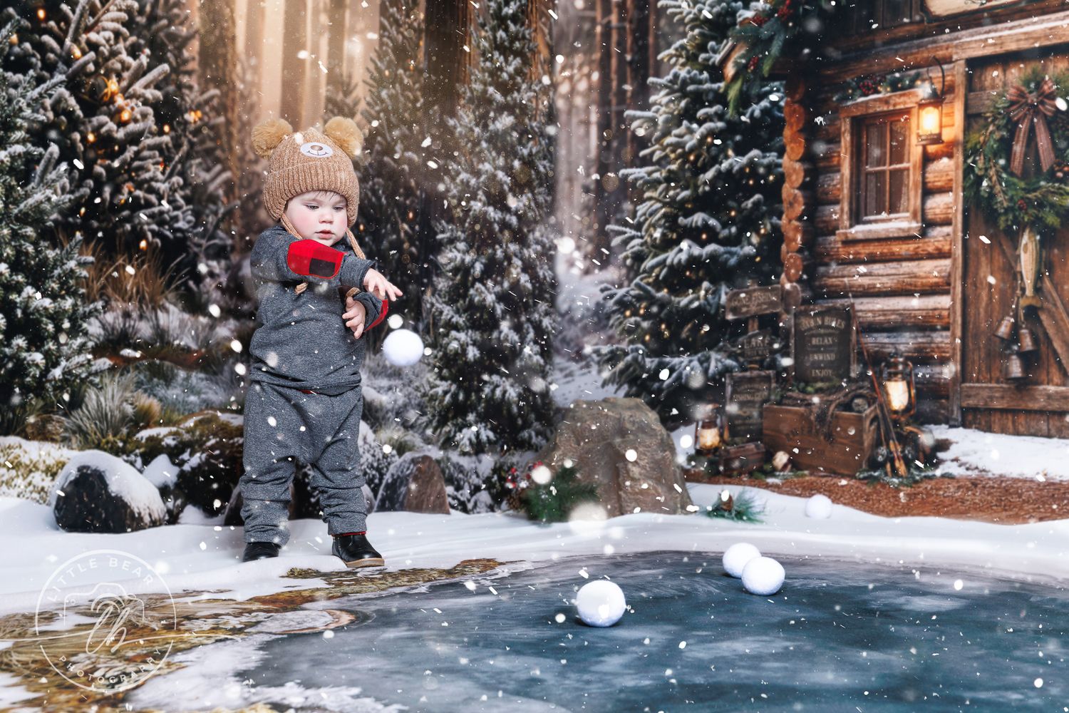 Toddler in winter snowsuit playing with snowballs near a cozy log cabin surrounded by snow-covered Christmas trees.