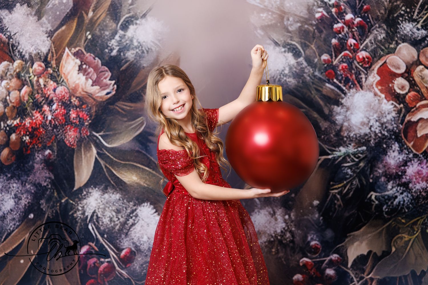 Smiling girl in red sequin dress holds giant Christmas ornament with festive floral backdrop.