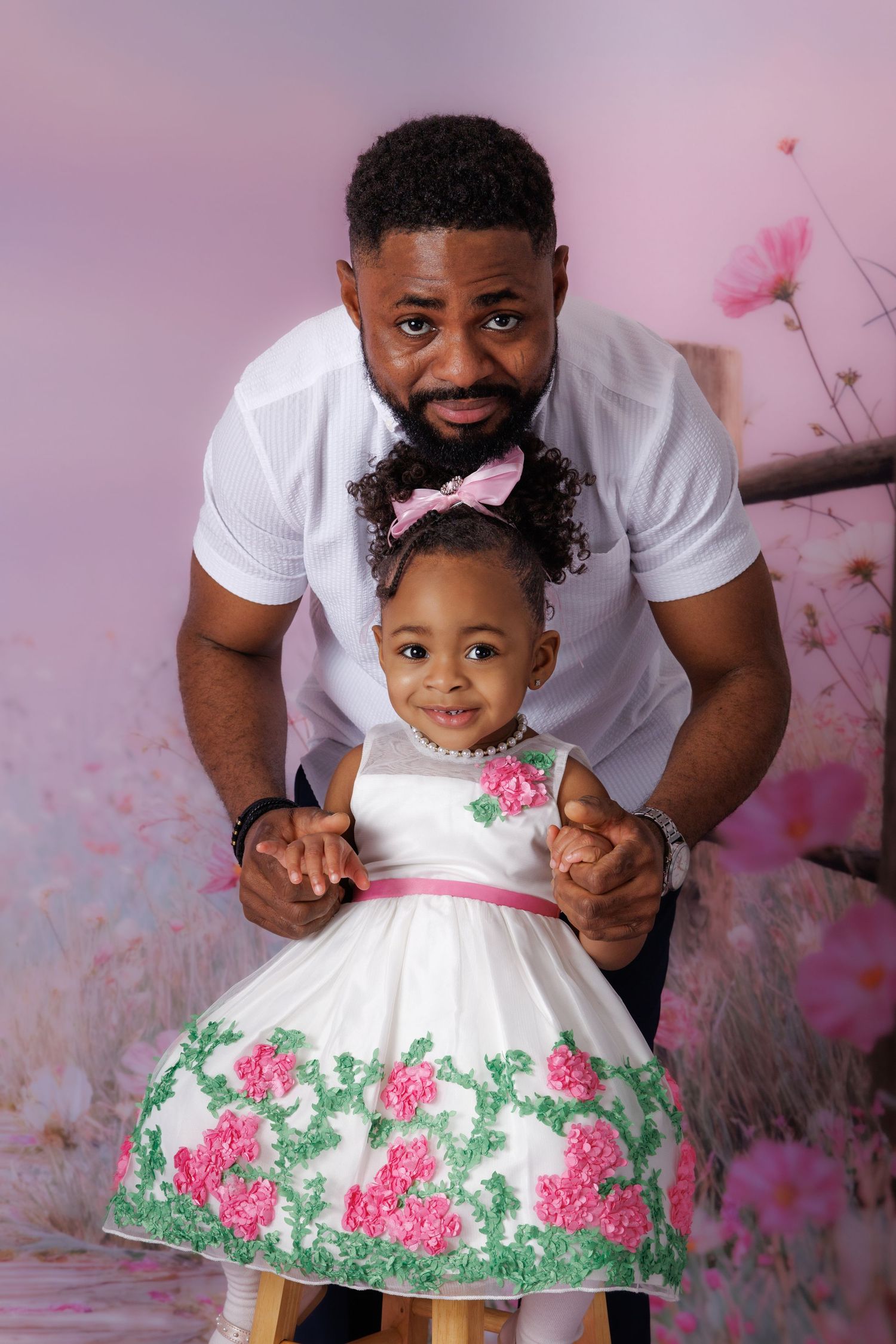 A father and young daughter pose together, she wears a white floral dress with pink bow, against a pink floral backdrop.