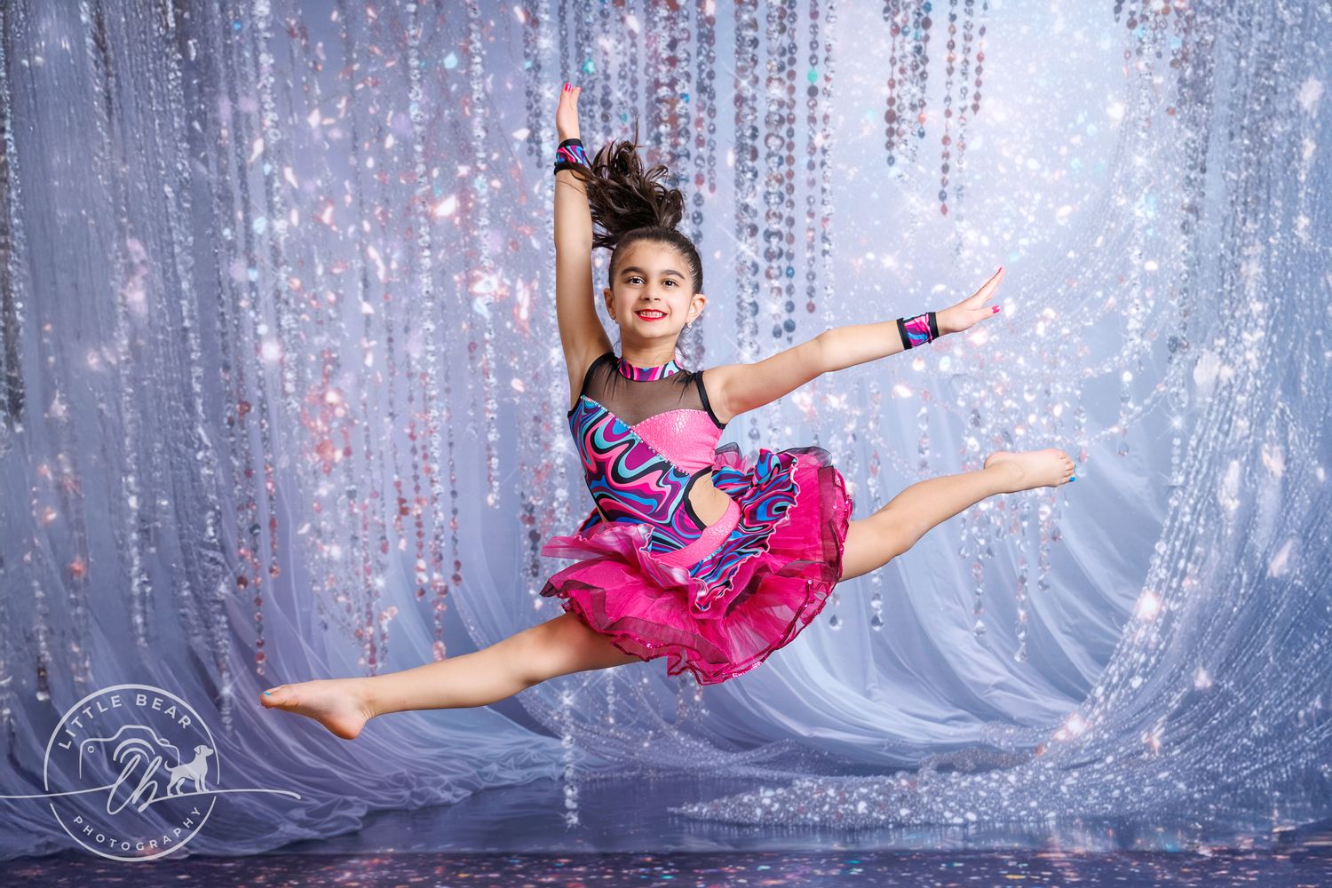 Young dancer in pink tutu performs a grand jeté split leap on sparkly stage.