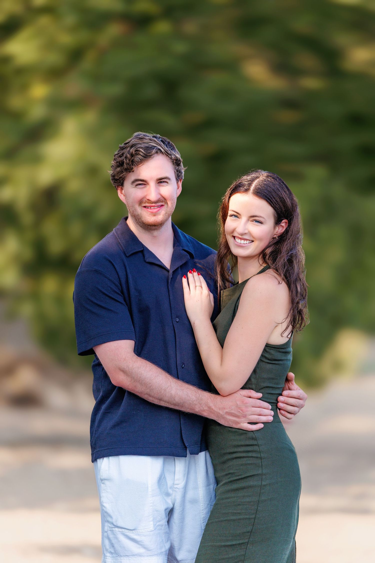 Smiling couple embracing outdoors, man in navy shirt and woman in olive green dress with bokeh background.