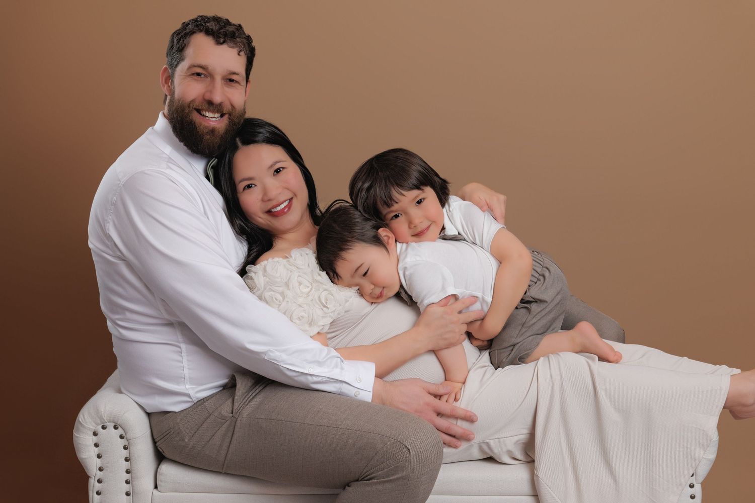Happy family of four with pregnant mother sitting together on white sofa against warm brown background.