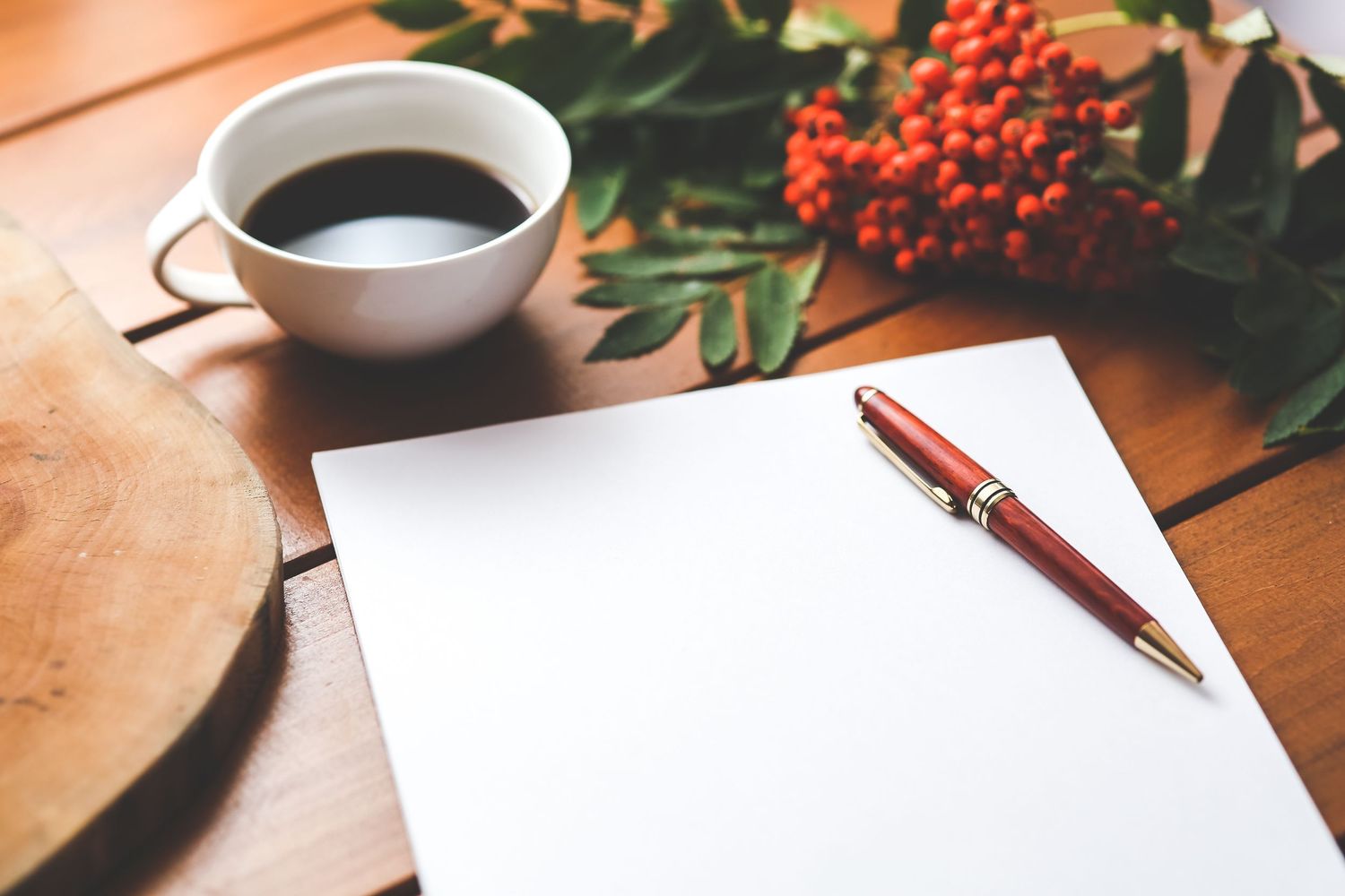 Blank white notepad with red pen beside black coffee cup and red berries on wooden table.