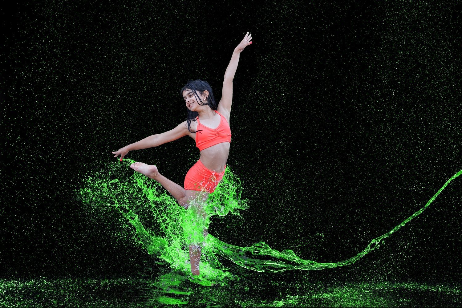 A dancer leaps gracefully amid vibrant green liquid splashes against a dark background, wearing a bright orange athletic outfit.