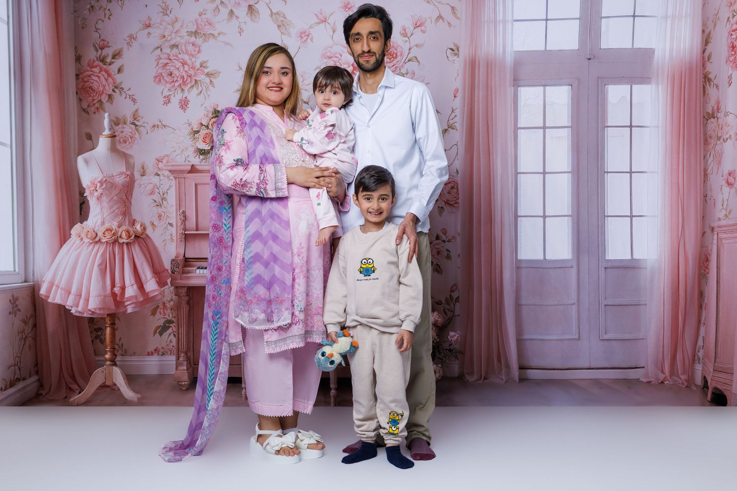 Family of four poses in pink floral studio with mother in purple tie-dye dress holding baby girl.
