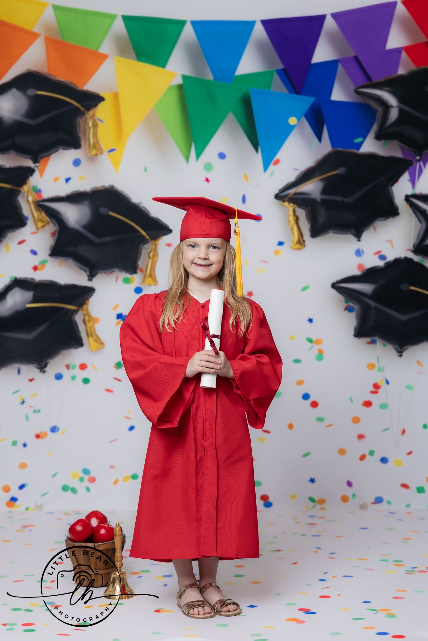 Young girl in red graduation cap and gown holds diploma, surrounded by mortarboard balloons and colorful confetti.