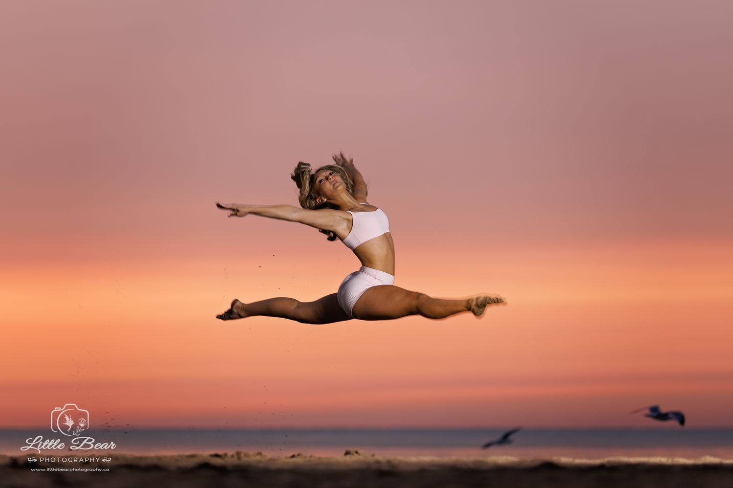 Dancer leaping gracefully at sunset on a beach, performing a split jump against a vibrant pink and orange sky.