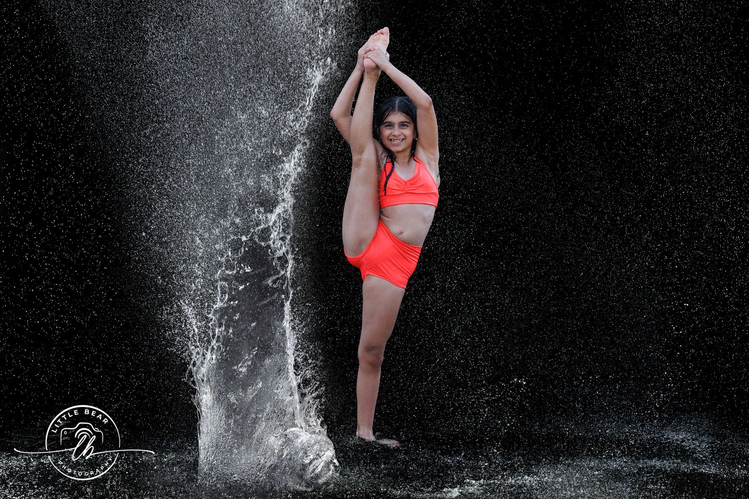 Woman in orange bikini performing a graceful dance pose under a waterfall splash against a dark background.