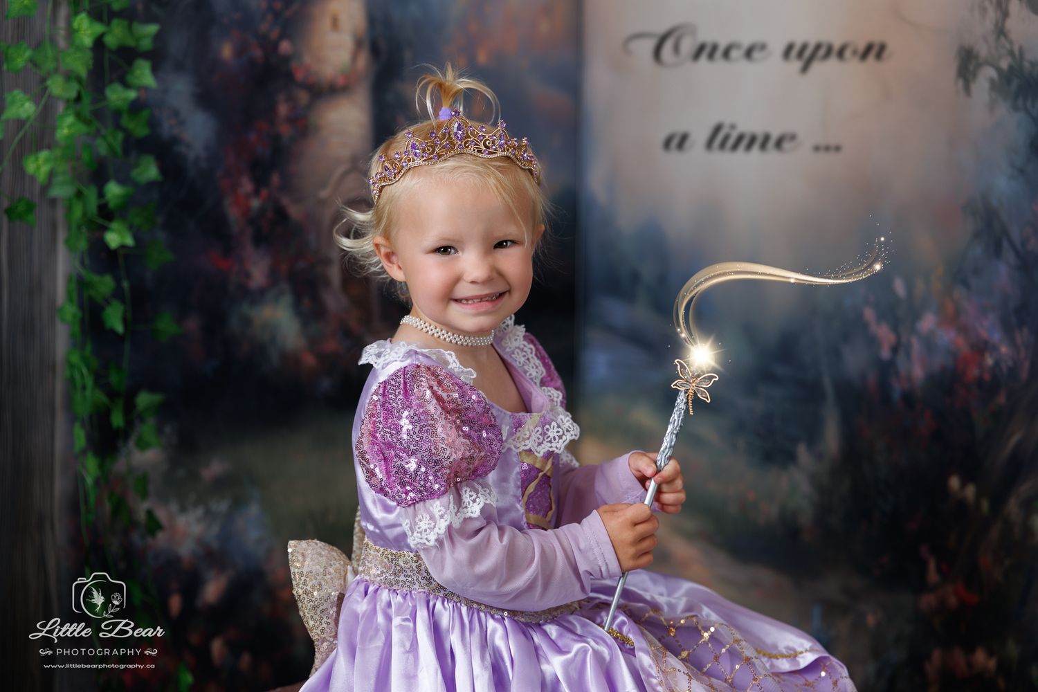 A smiling toddler girl in a purple princess dress and tiara holds a magic wand in a fairy tale photo setting.