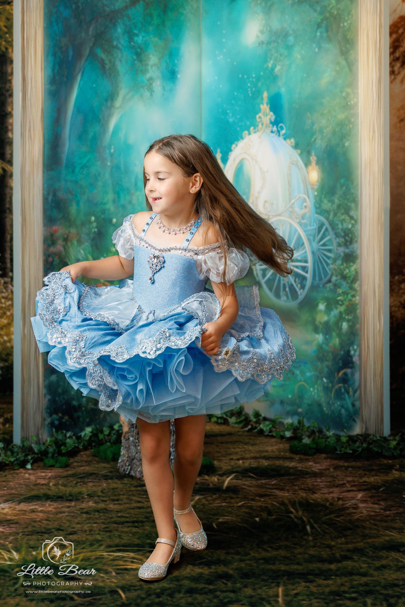 Young girl in blue Cinderella dress twirling in front of enchanted forest backdrop with carriage.