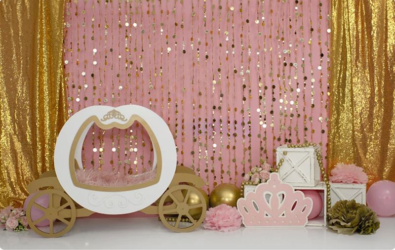 Pink and gold princess carriage cake smash backdrop with sequin curtains, crown decorations, and floral accents.