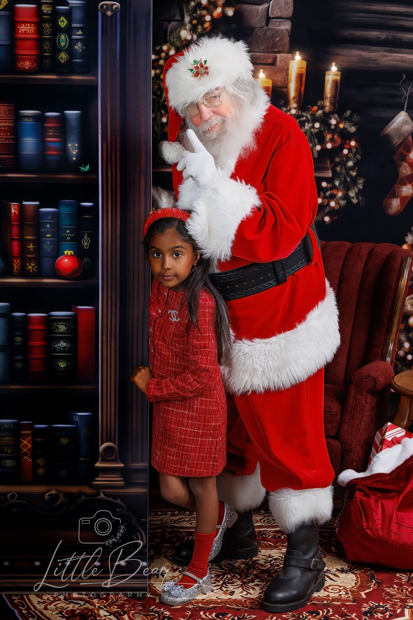 Young girl in red dress poses with Santa Claus in festive holiday portrait setting with bookshelf and Christmas decorations.