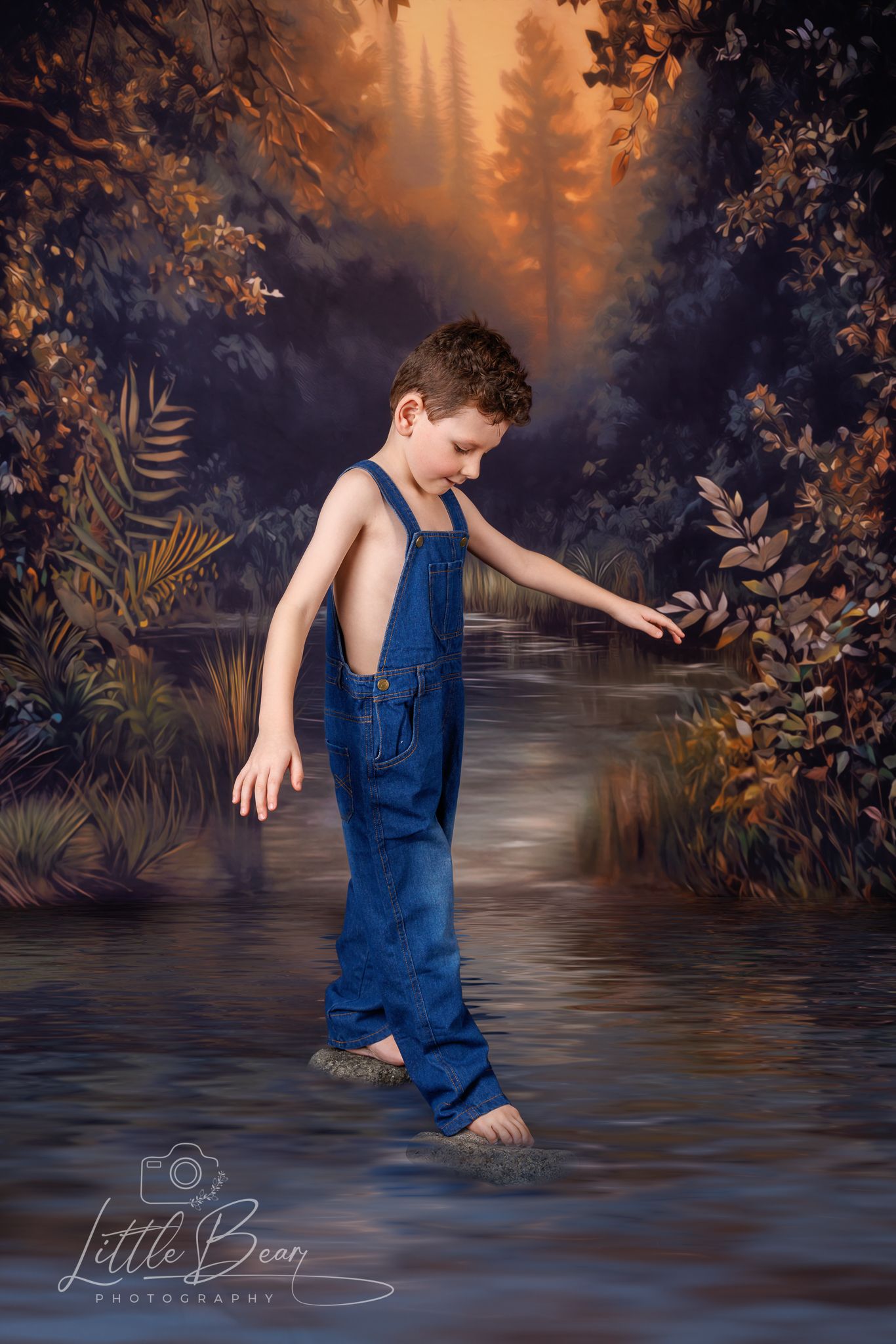Boy in blue denim overalls wades through water in a golden autumn forest photography backdrop.