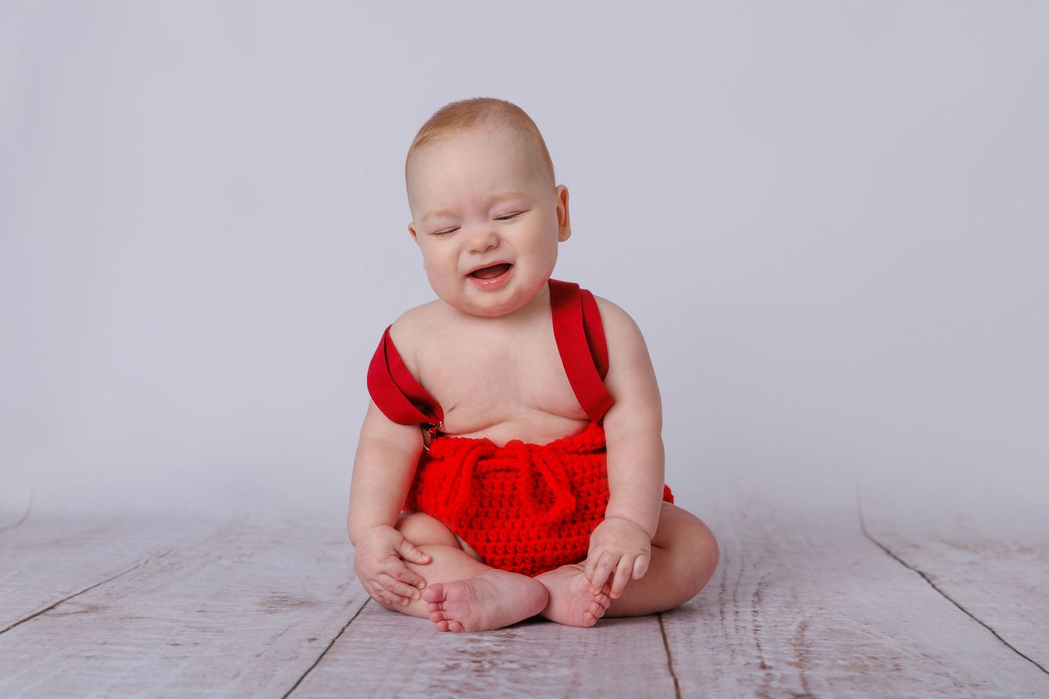 Adorable baby in red romper sitting on white wood floor, eyes closed, smiling joyfully.