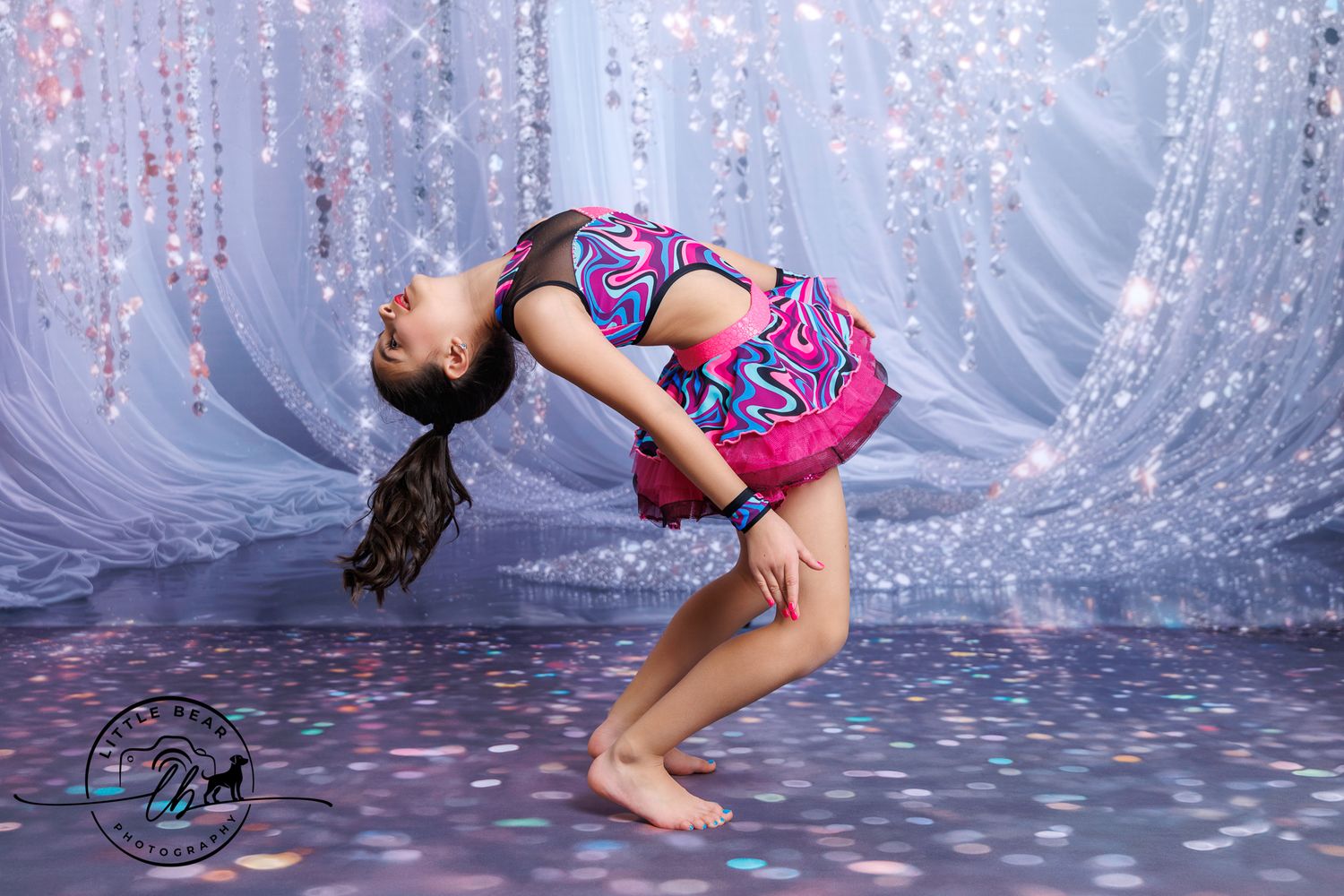 Young dancer in colorful costume performs a deep backbend on a glittery stage.