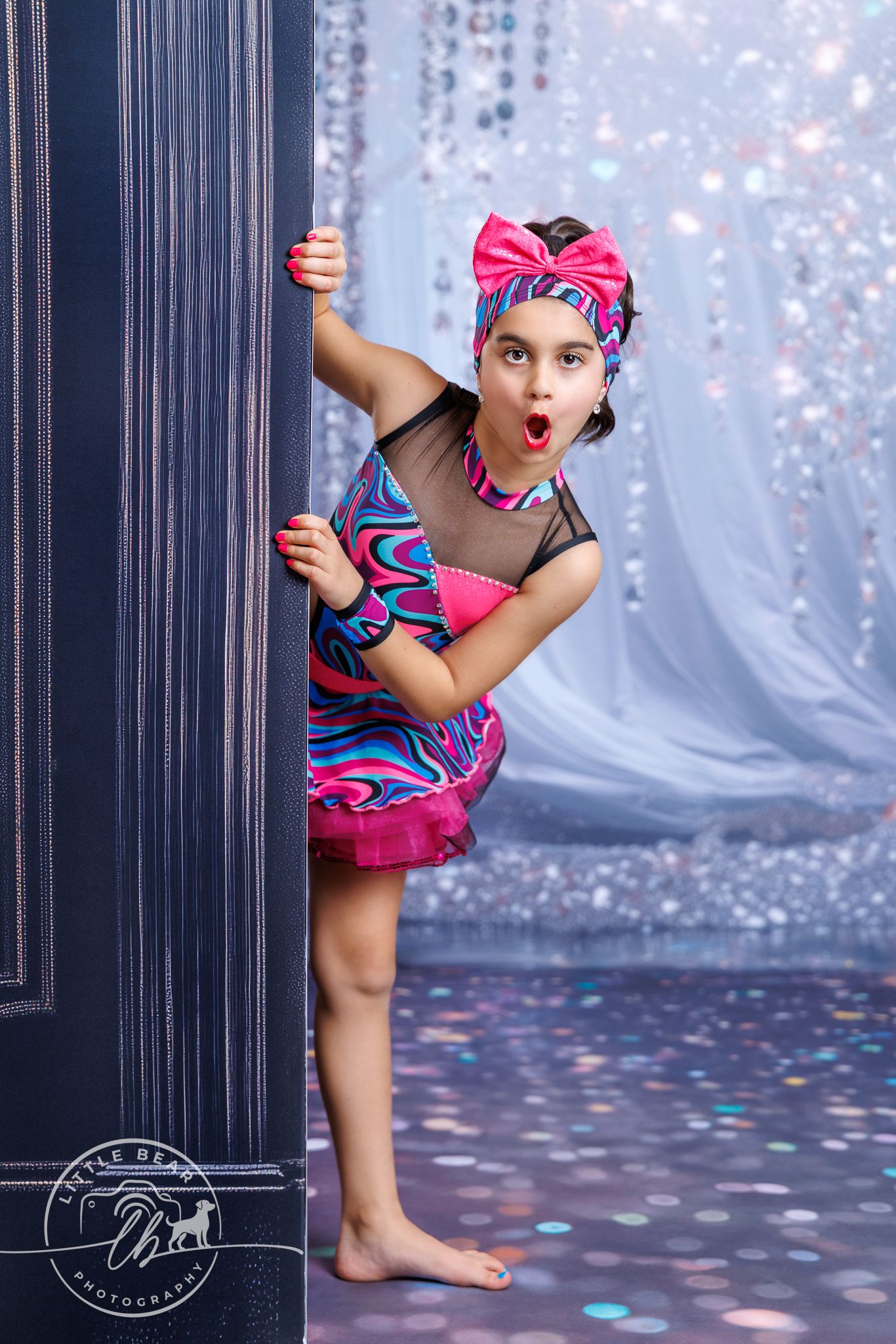 Young girl in colorful pink dance costume peeks playfully around a dark column with a surprised expression on her face.