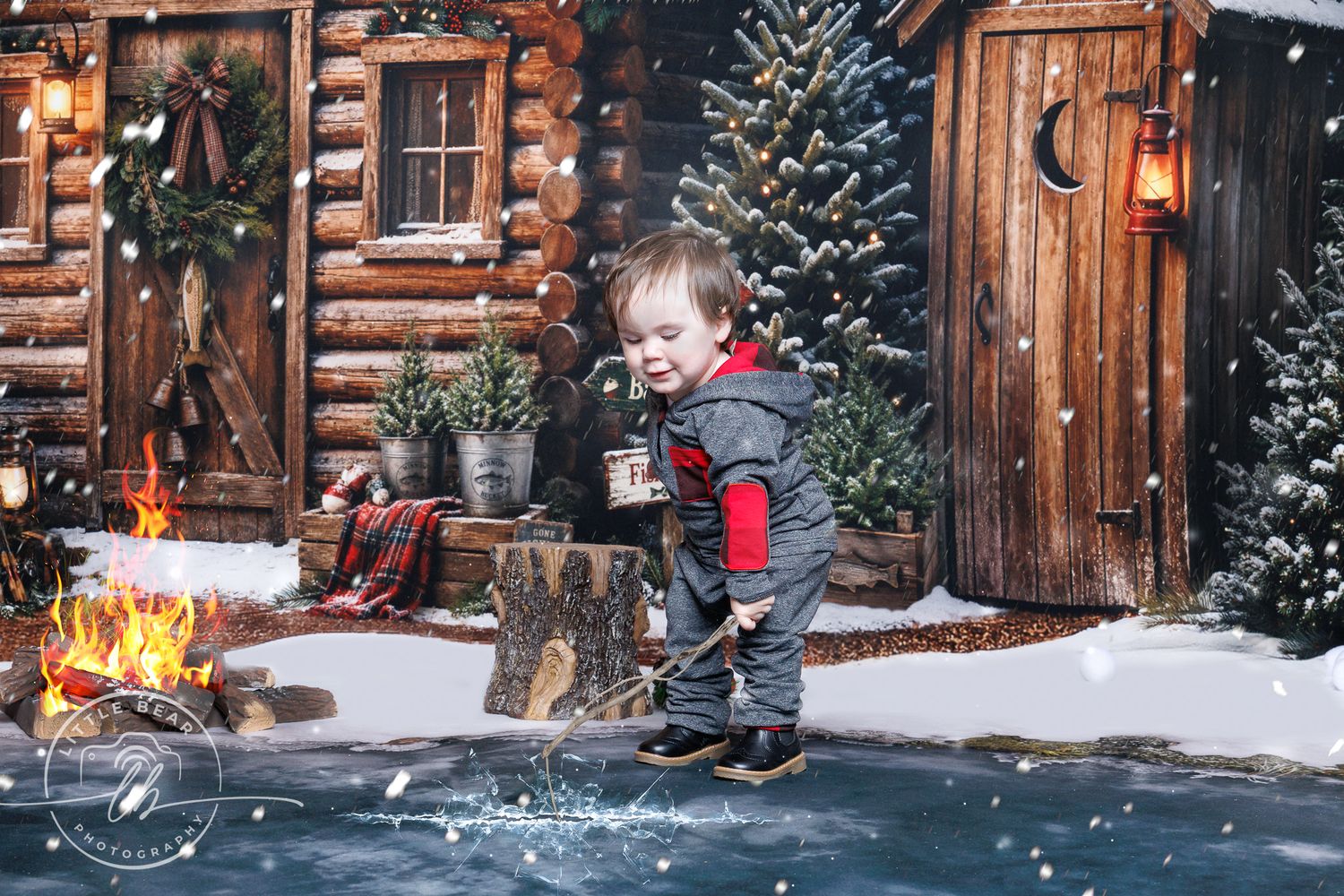 A laughing toddler boy plays in snow beside a rustic log cabin with Christmas decorations and a campfire.