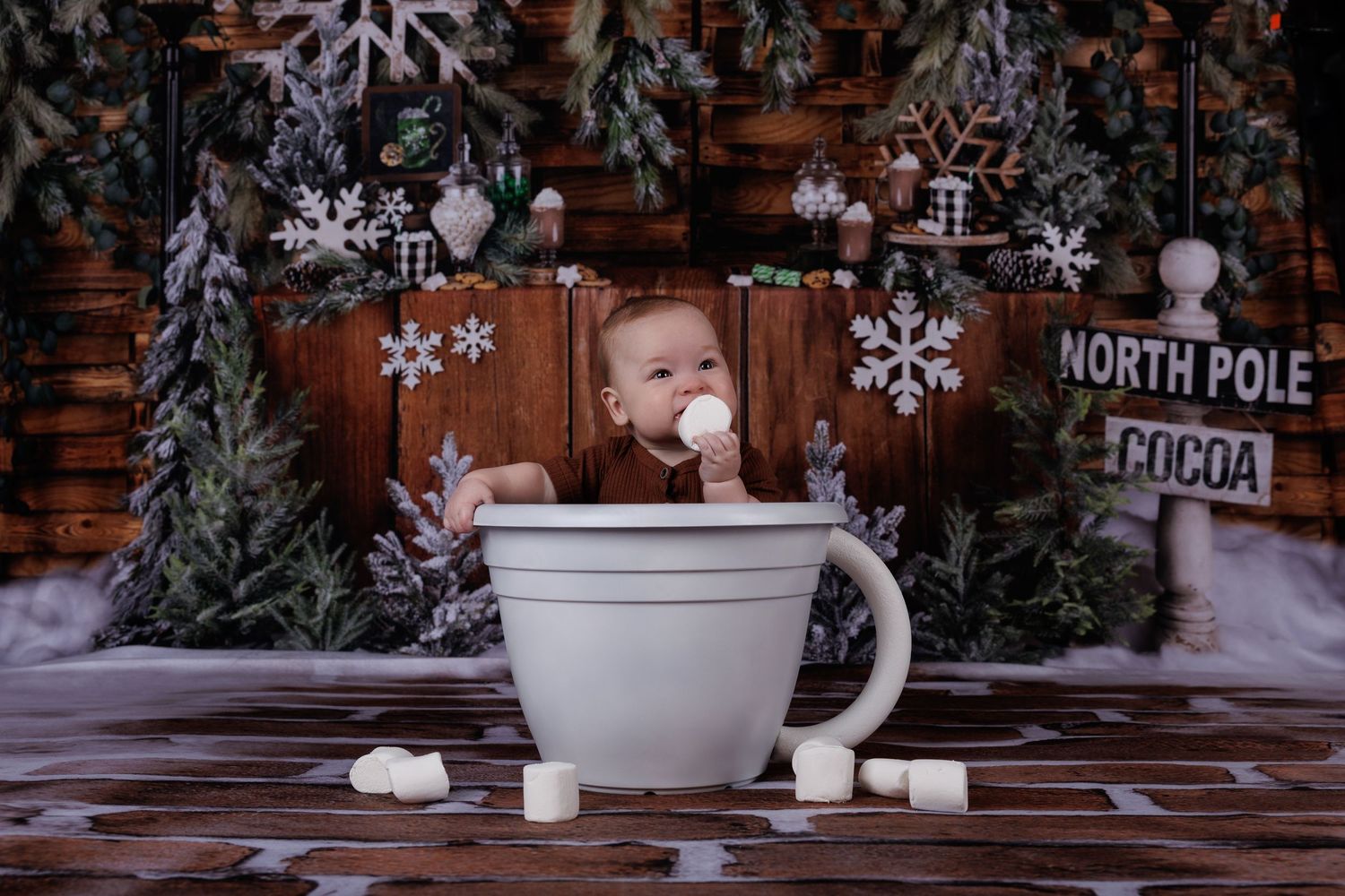 Cute baby sits inside oversized mug prop in a festive North Pole hot cocoa Christmas scene.