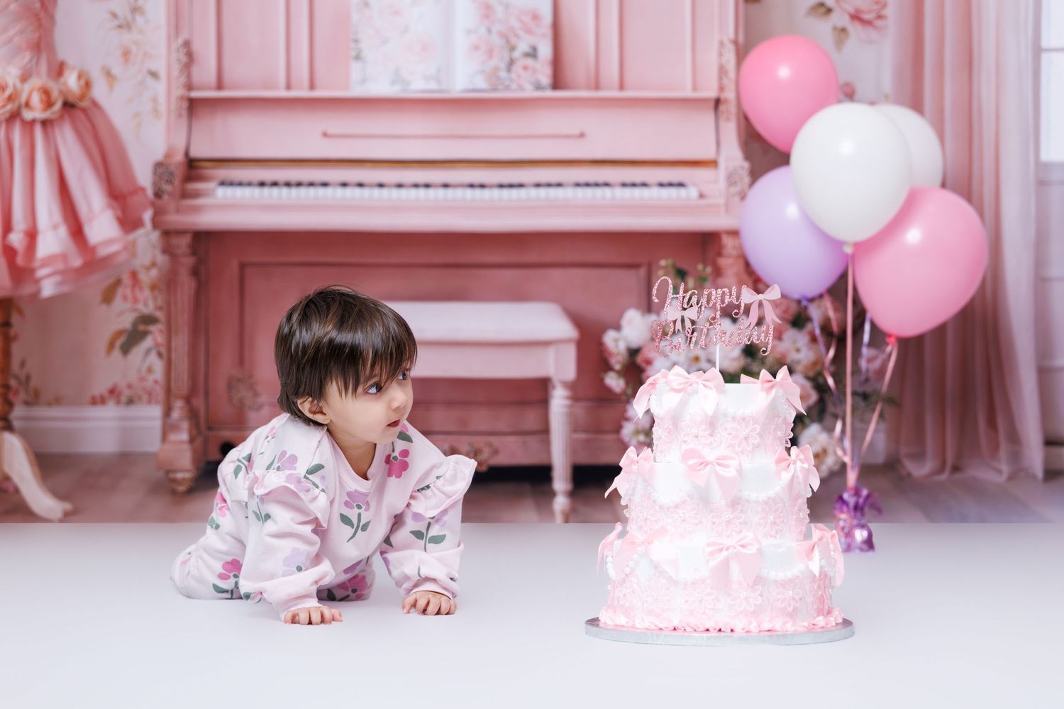 A baby girl in a floral outfit crawls toward a pink tiered birthday cake with balloons.