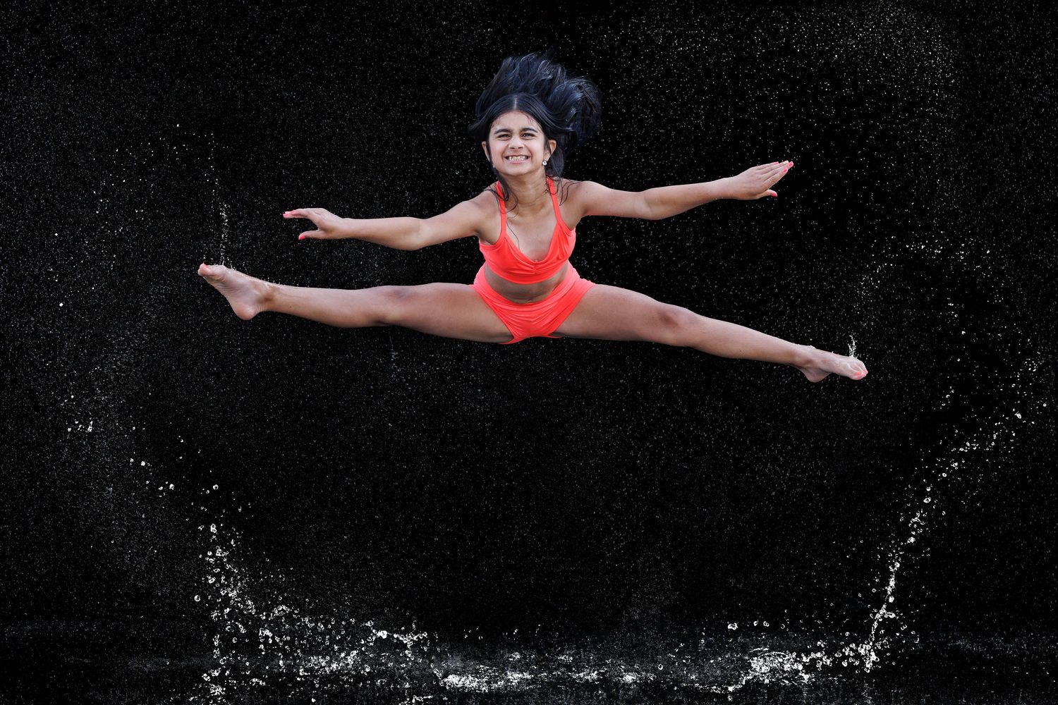 Woman in red swimsuit performing a mid-air split leap with water splashes on black background.
