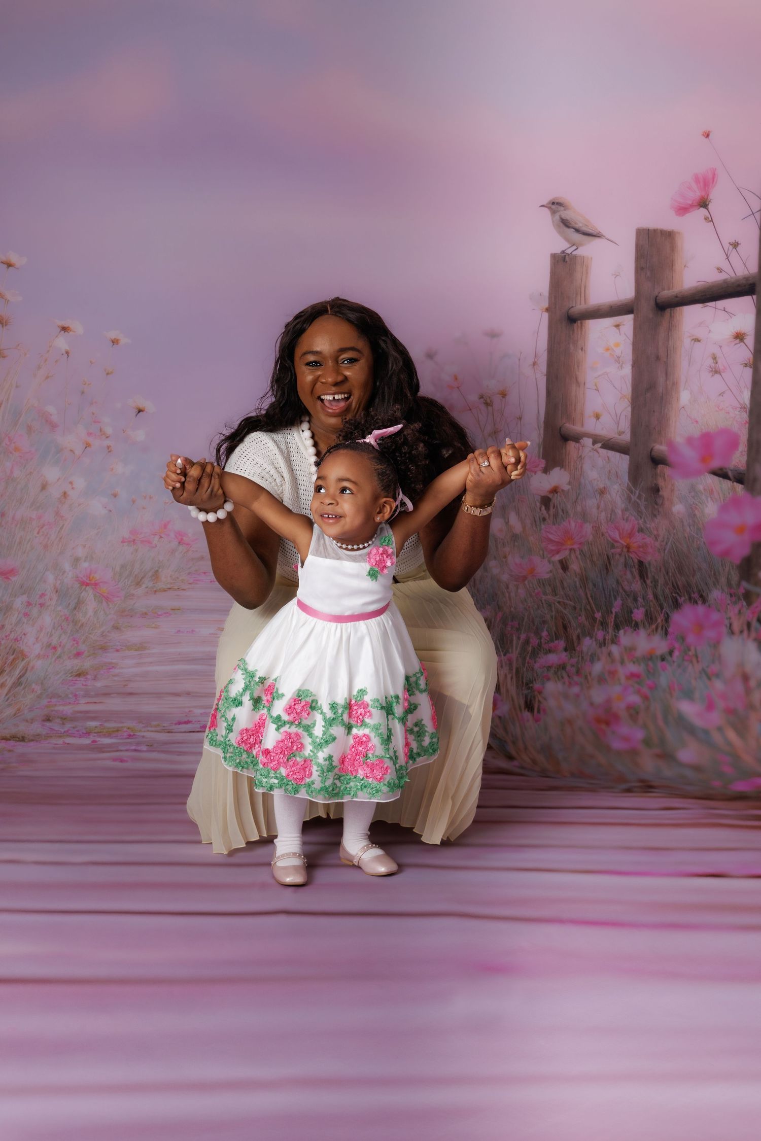 A mother helps her baby daughter stand in matching white dresses against a pink floral garden backdrop.