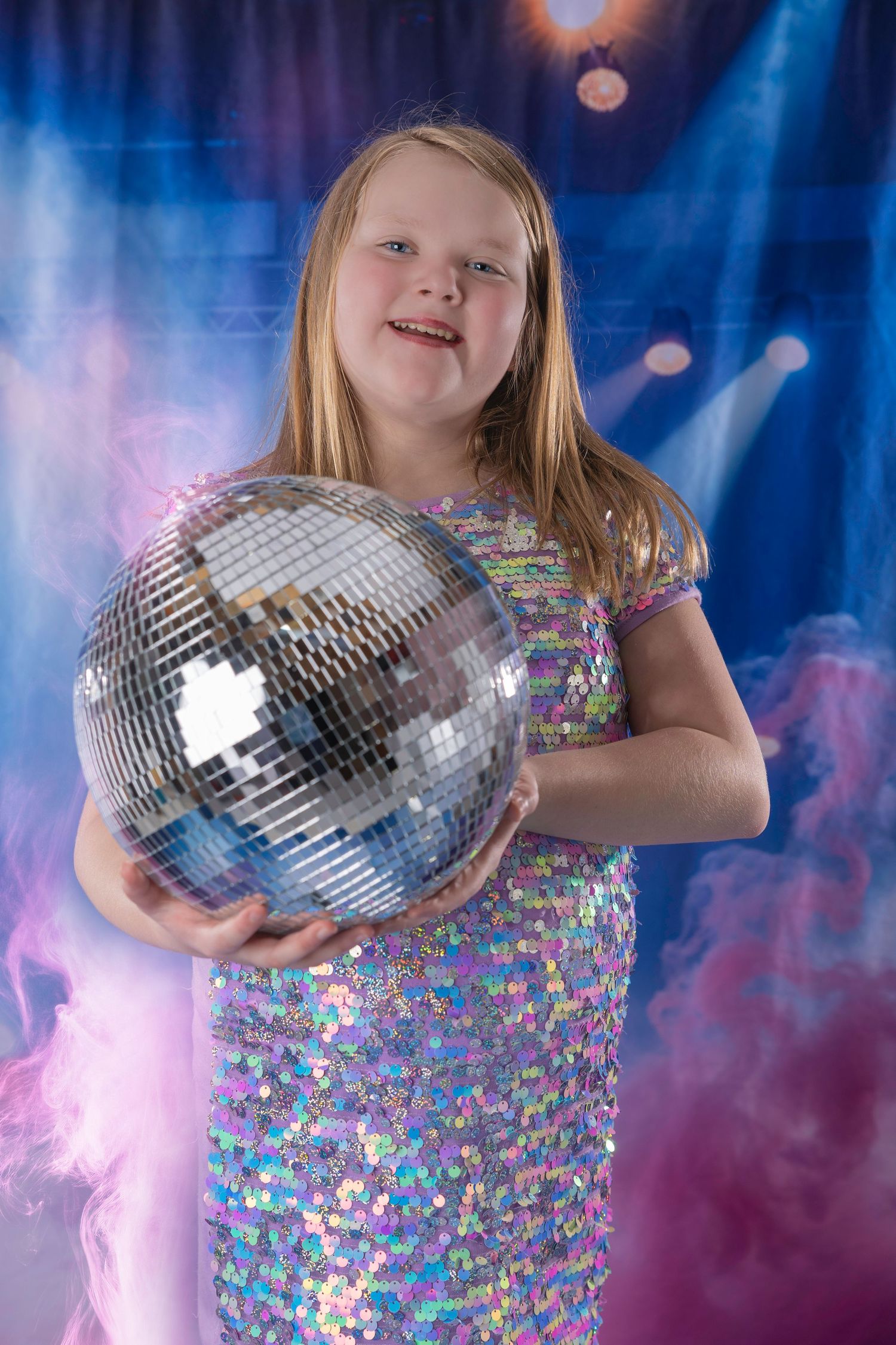 Young girl in sequin dress holding a large disco mirror ball on a colorful stage with smoke effects.