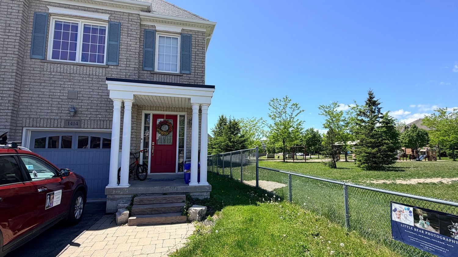 A brick two-story home with white columns, a red front door wreath, and a red car in the driveway on a sunny day.