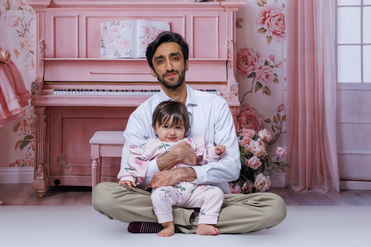 A man holds a baby girl on his lap while seated in a pink floral photography studio.