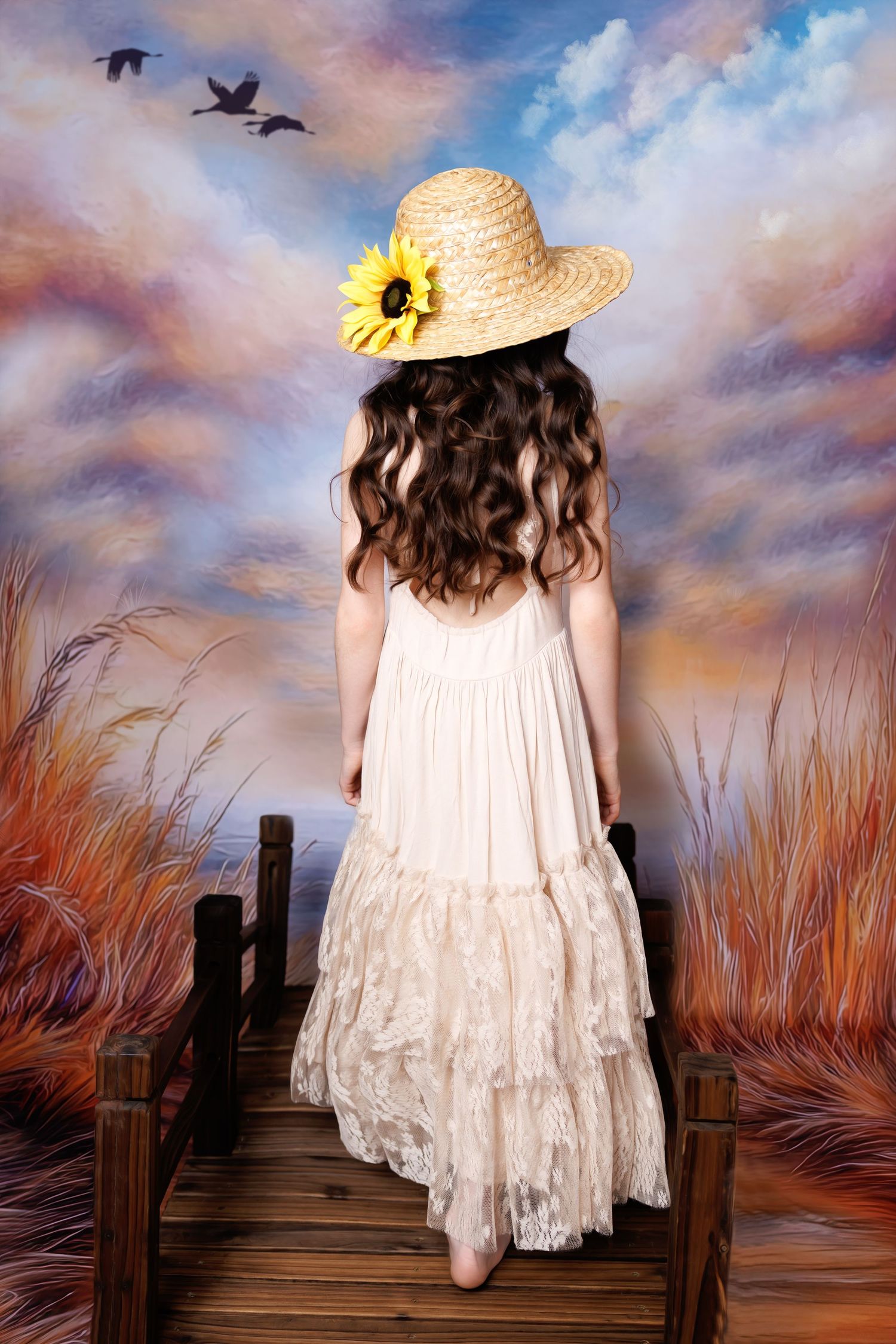 A young girl in a cream lace dress and sunflower straw hat stands with her back turned on a wooden bridge.