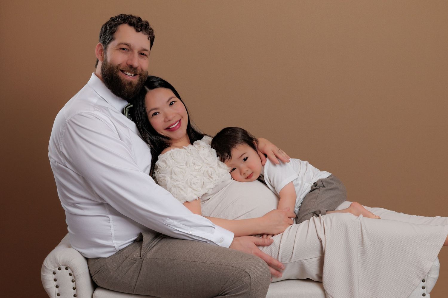 Family maternity portrait with couple and young child sitting together on a white chair against brown backdrop.