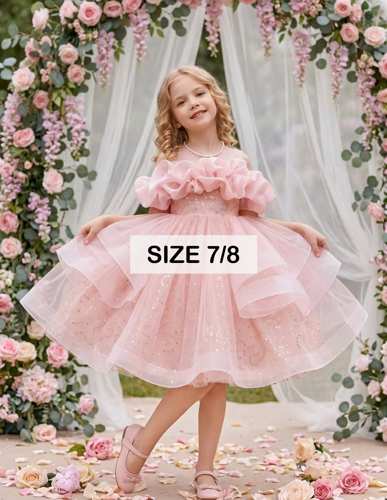 Girl wearing a pink sparkly tiered tulle dress size 7/8 with ruffled sleeves, posing in front of floral arch.