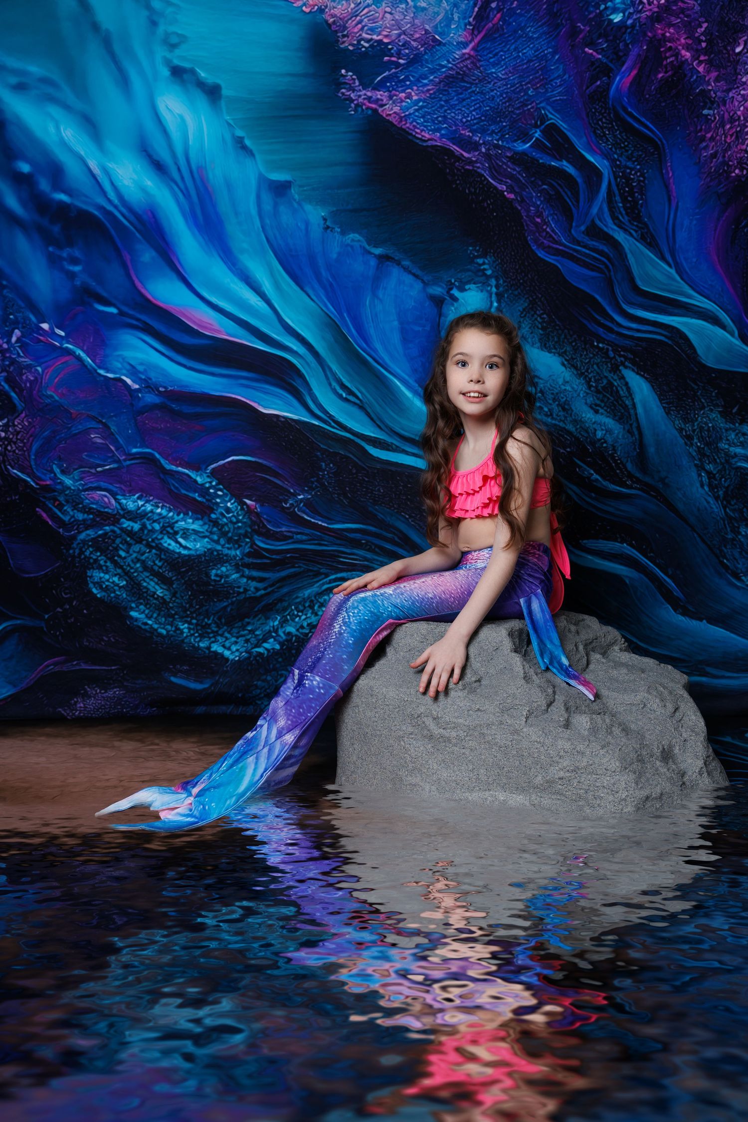 Smiling girl in pink and purple mermaid tail costume sits on rock with blue wave background.