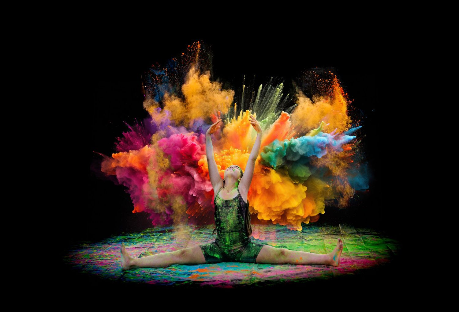 Dancer kneels surrounded by massive multicolored powder explosion of orange, yellow and blue hues.