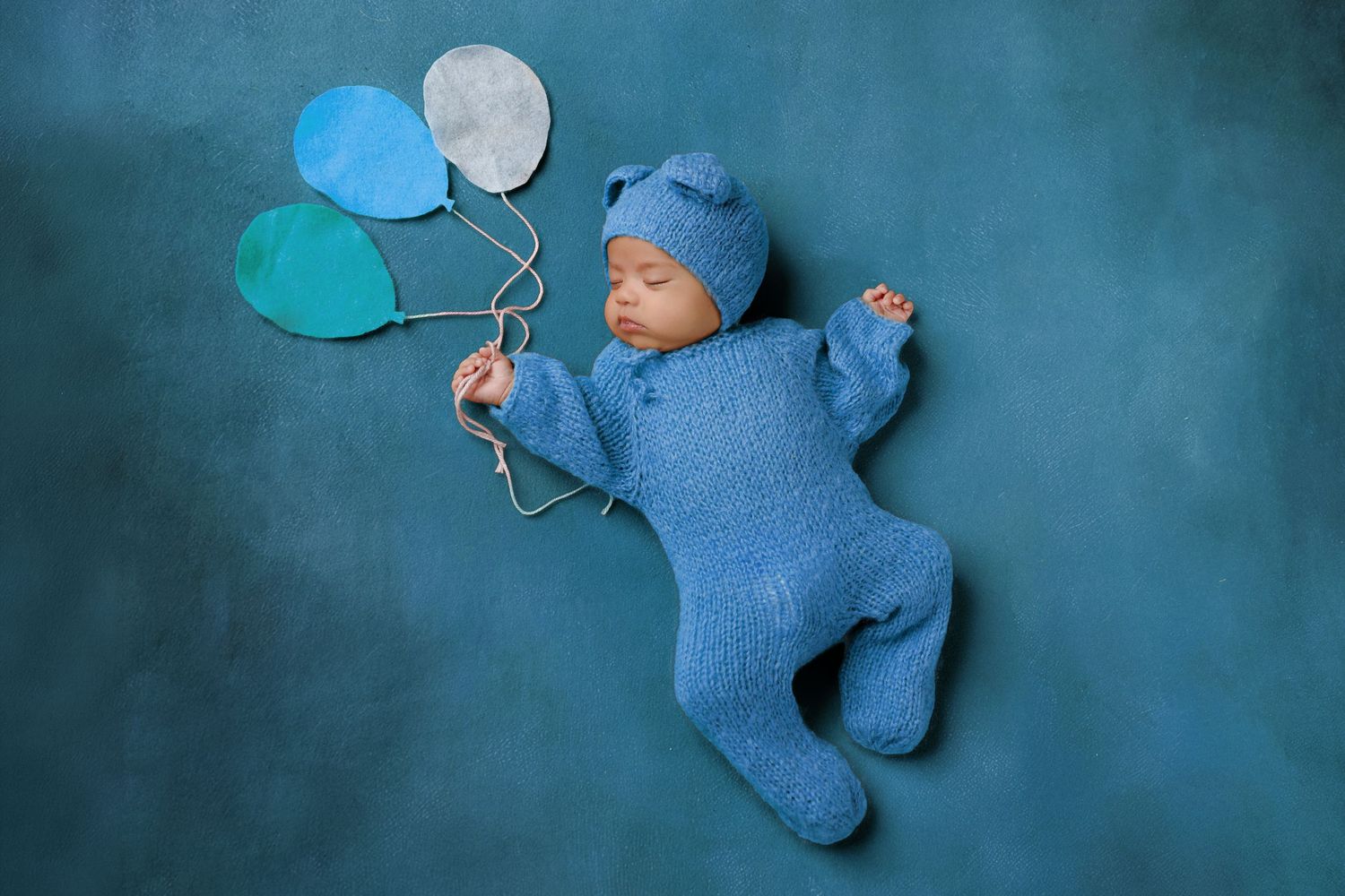 Newborn baby in blue knit outfit holds colorful paper balloons on teal background.