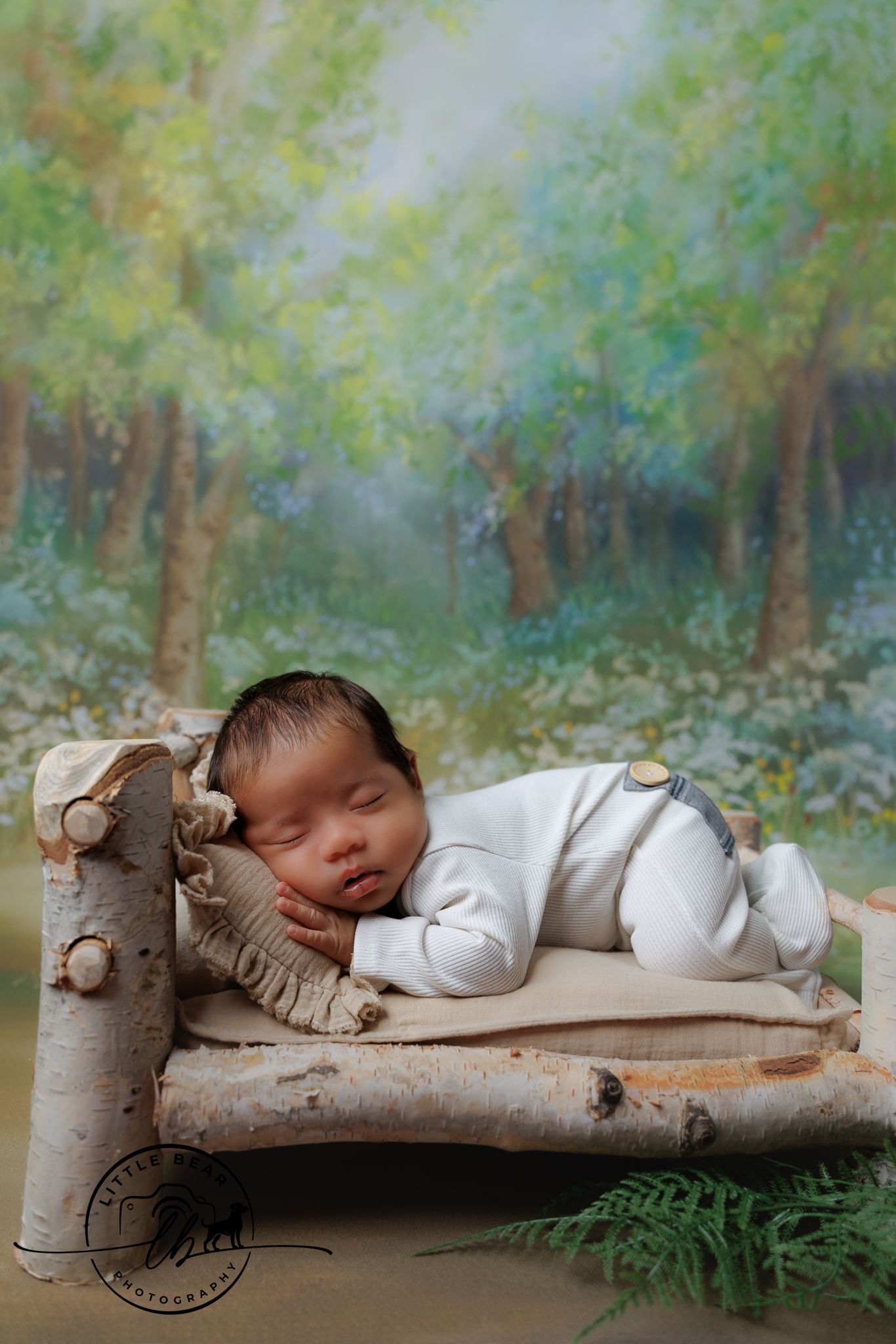 Sleeping newborn in white outfit resting peacefully on birch wood bed with lush forest painted backdrop.