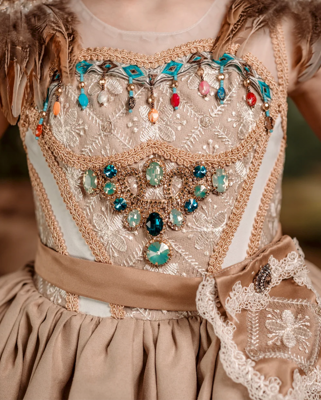 Ornate beige gown with teal, red, and green gemstone embellishments, gold trim, lace details, and satin ribbon waist sash.