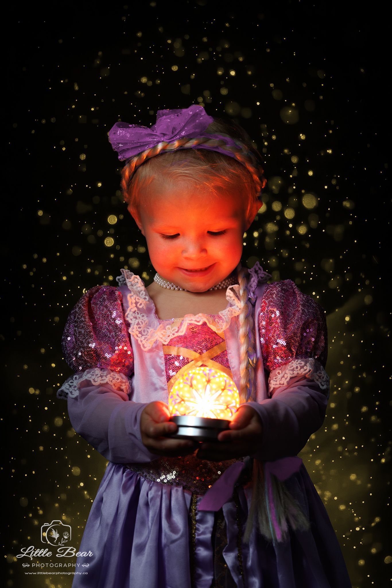 A toddler girl in a purple Rapunzel princess dress holds a glowing orb, illuminated by magical golden bokeh lights.