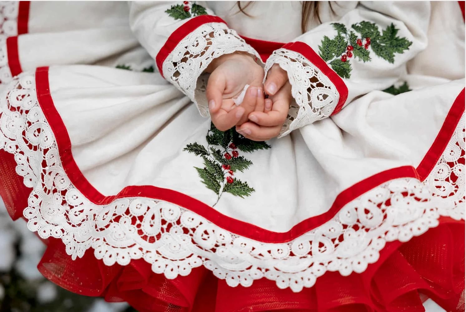Child in festive white dress with red trim, lace details, and holly embroidery, hands clasped together.