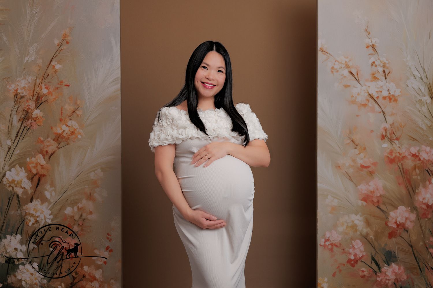 A smiling pregnant woman in a white dress poses gracefully before a soft floral beige studio backdrop.