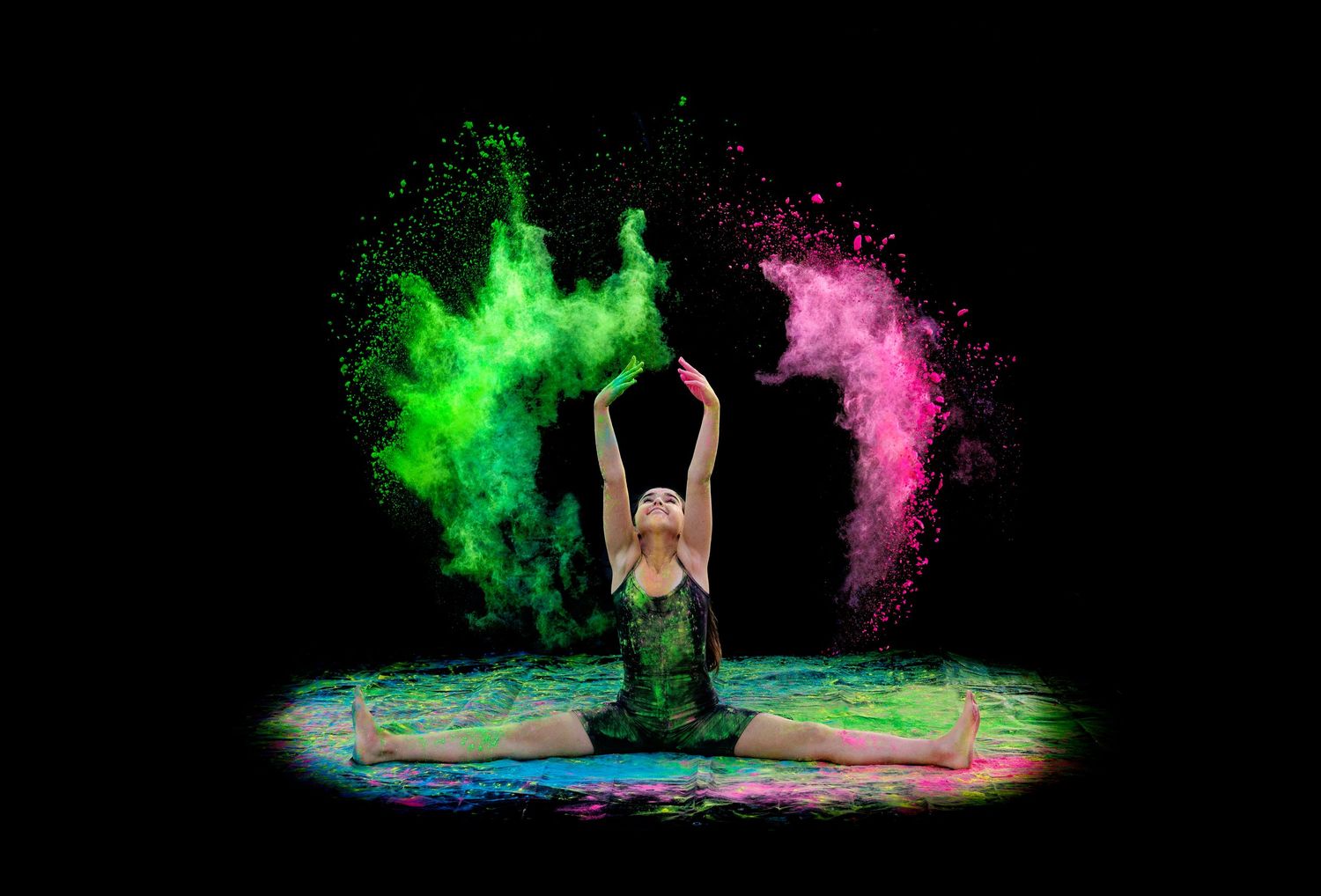 Gymnast in green leotard performing splits while tossing colorful powder against a dramatic black background.
