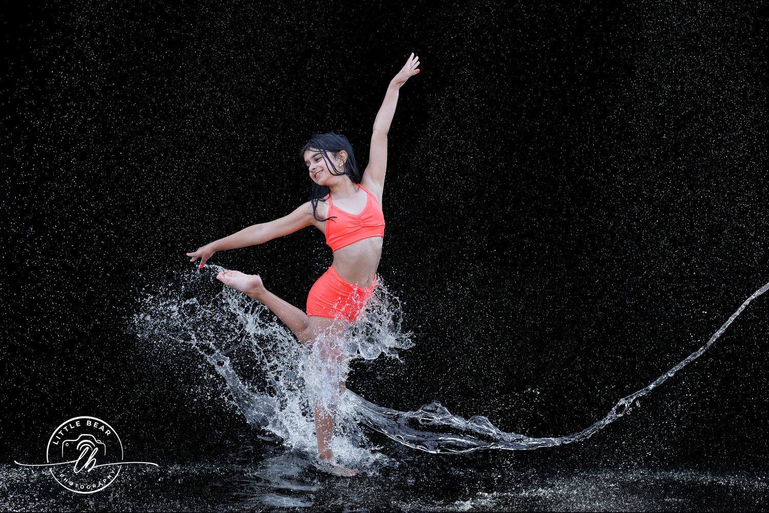 Dancer in orange outfit leaping gracefully through water splashes against a dark background.