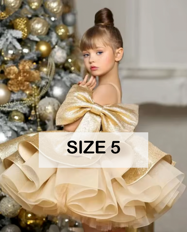 Girl in size 5 gold glitter ruffle ball gown dress with bow, posed near Christmas tree with ornaments.