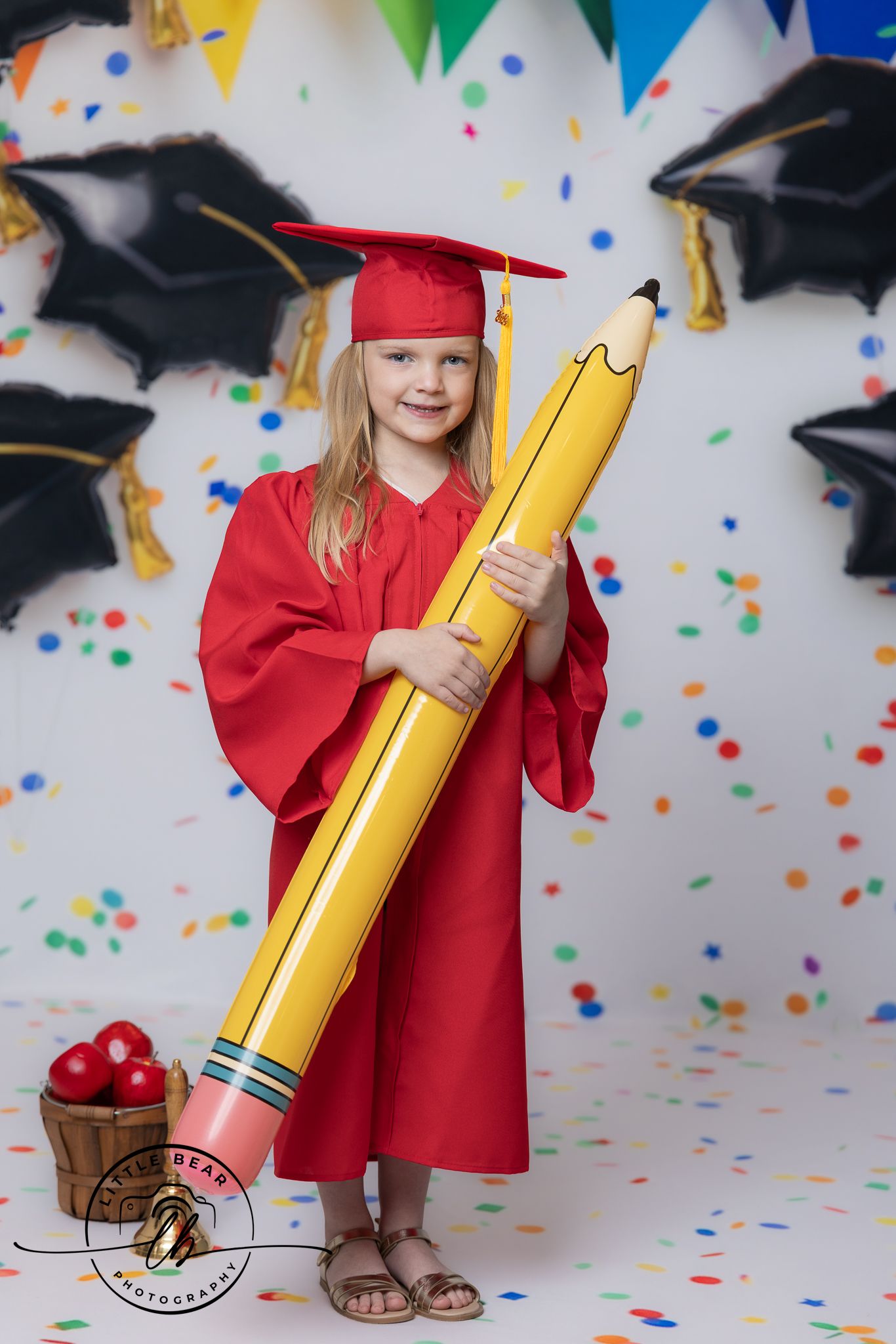 Young girl in red graduation cap and gown holding a giant yellow pencil prop with balloons and confetti backdrop.