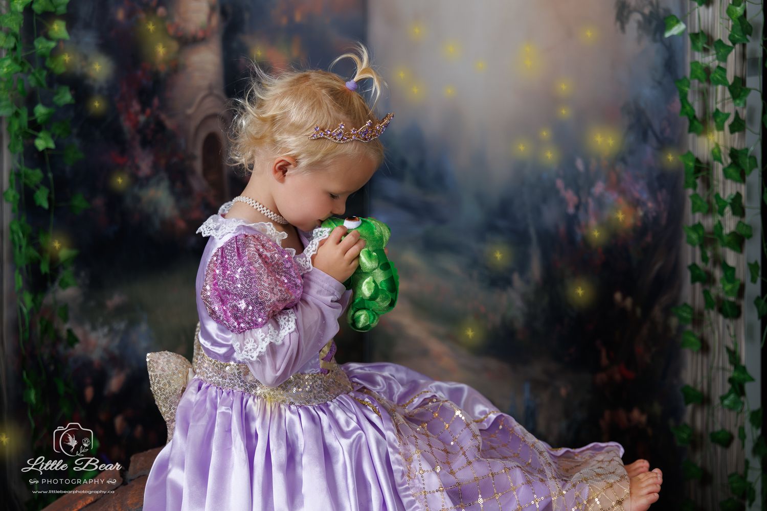 Toddler girl in purple Rapunzel princess dress holding green frog toy in enchanted forest photo backdrop.