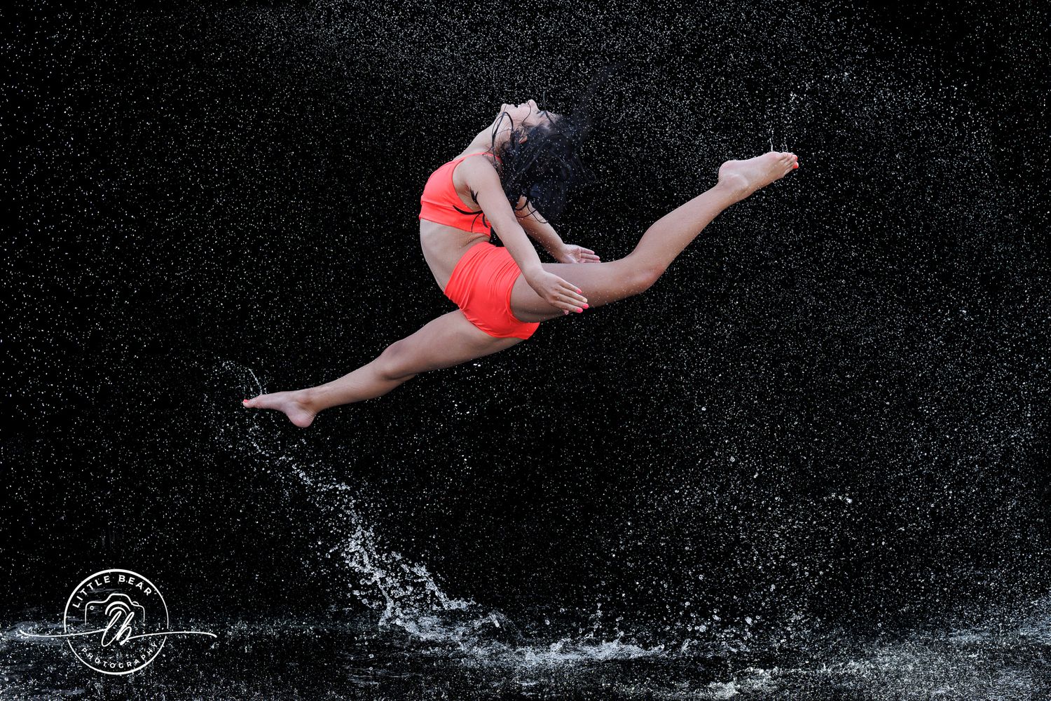 Dancer in red outfit leaping gracefully through water splashes against a dark background.