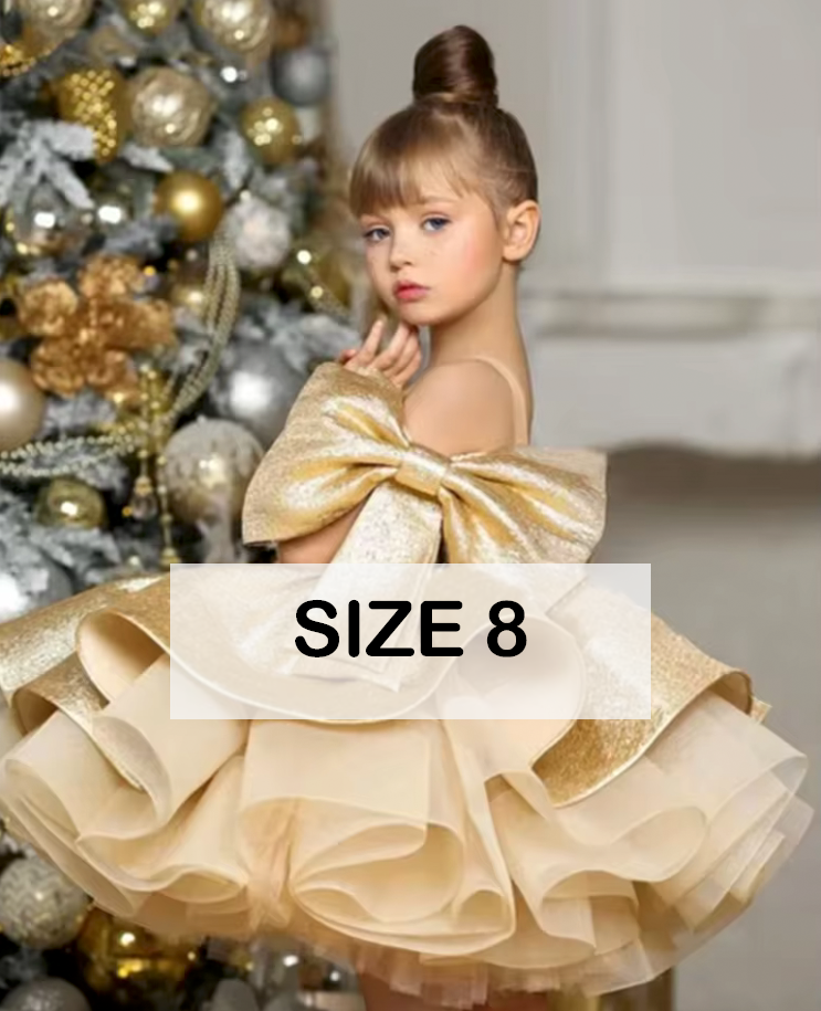 Girl in size 8 gold ruffled pageant dress with bow, posing near Christmas tree with ornaments.