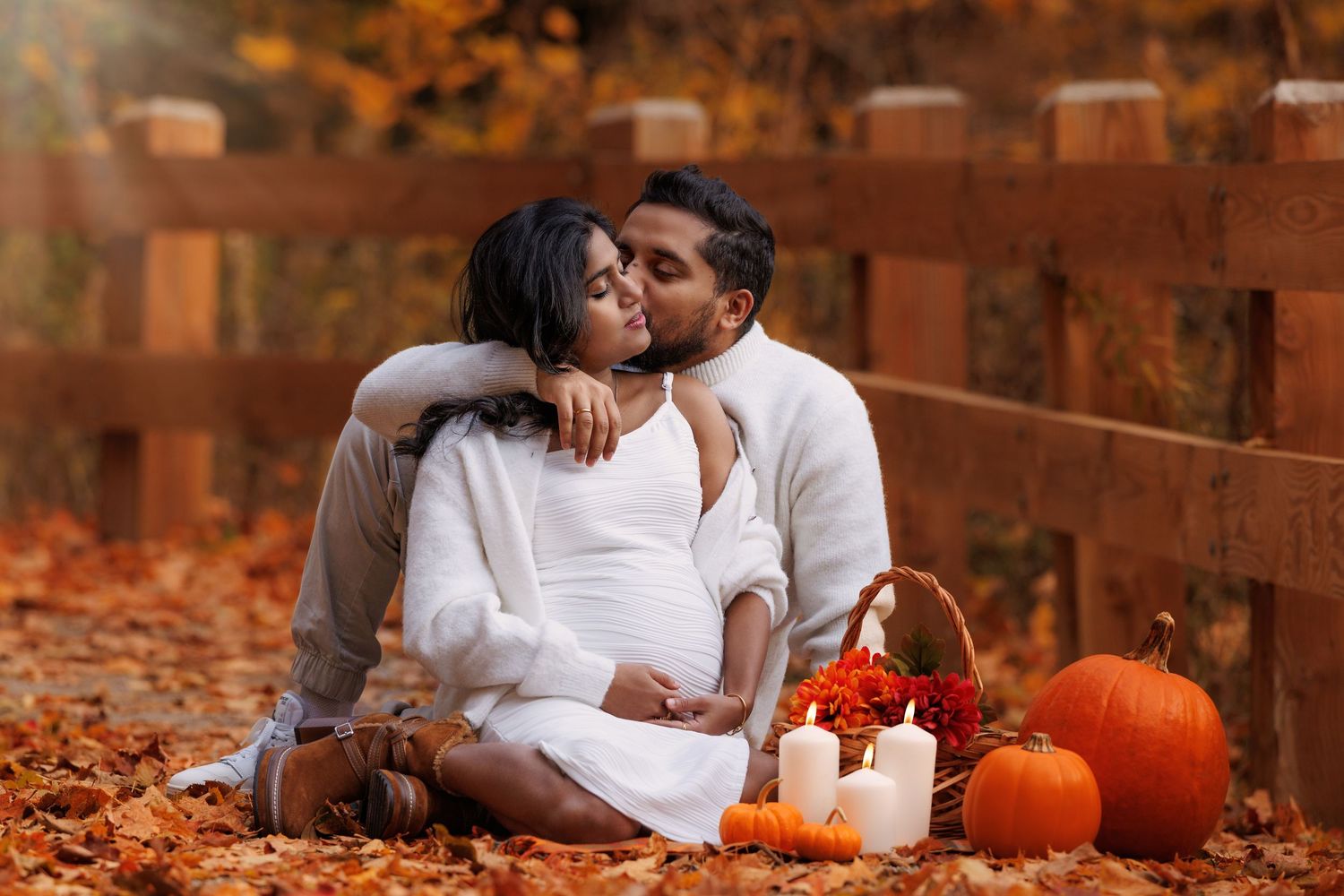 Pregnant couple sharing a kiss outdoors in autumn leaves with pumpkins and candles by a wooden fence.