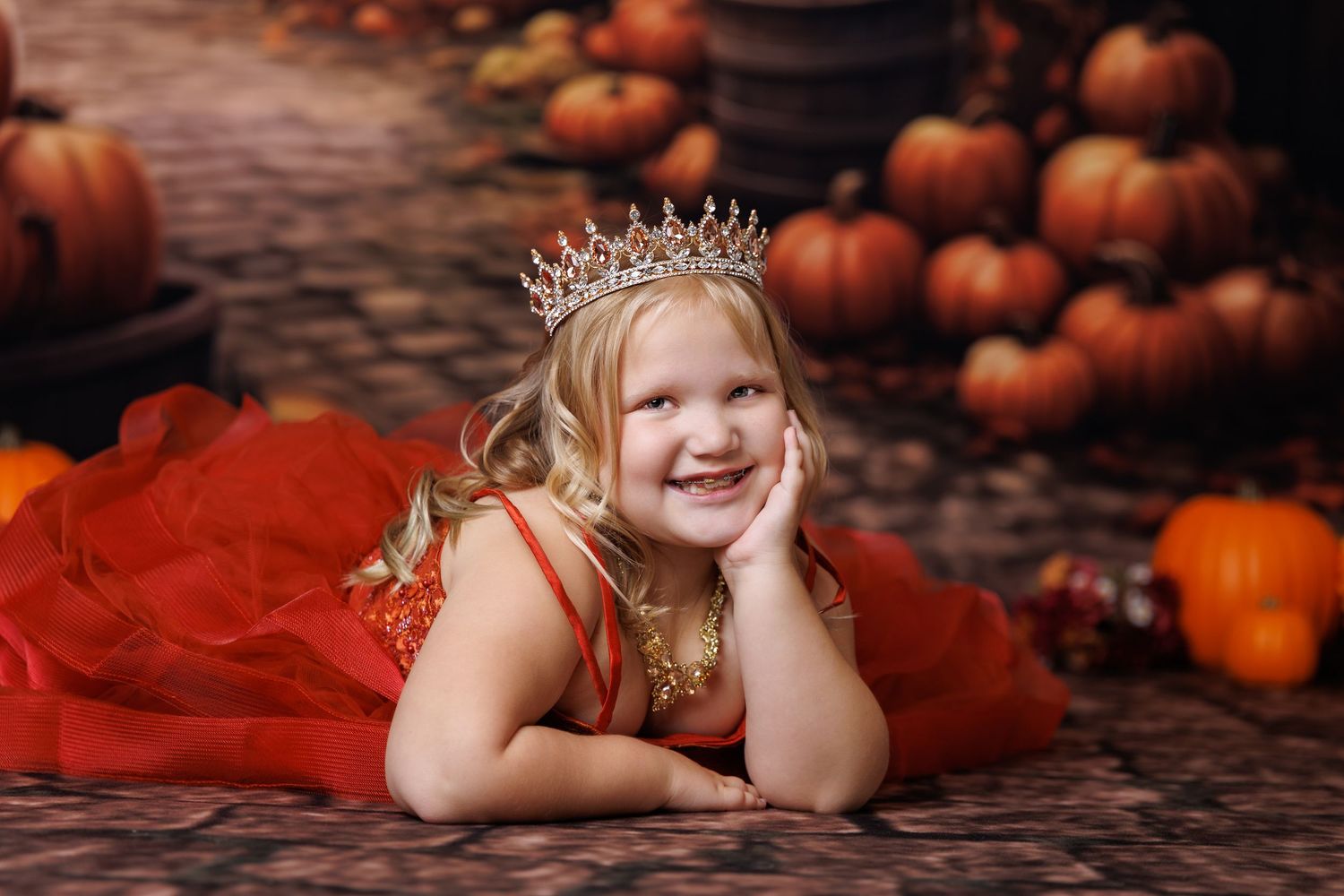 A smiling girl in a red dress and crown poses among pumpkins in a festive fall portrait.