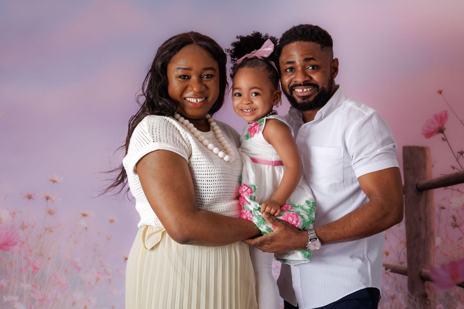 A happy family of three poses together, parents smiling warmly while holding their young daughter in a pink floral studio setting.