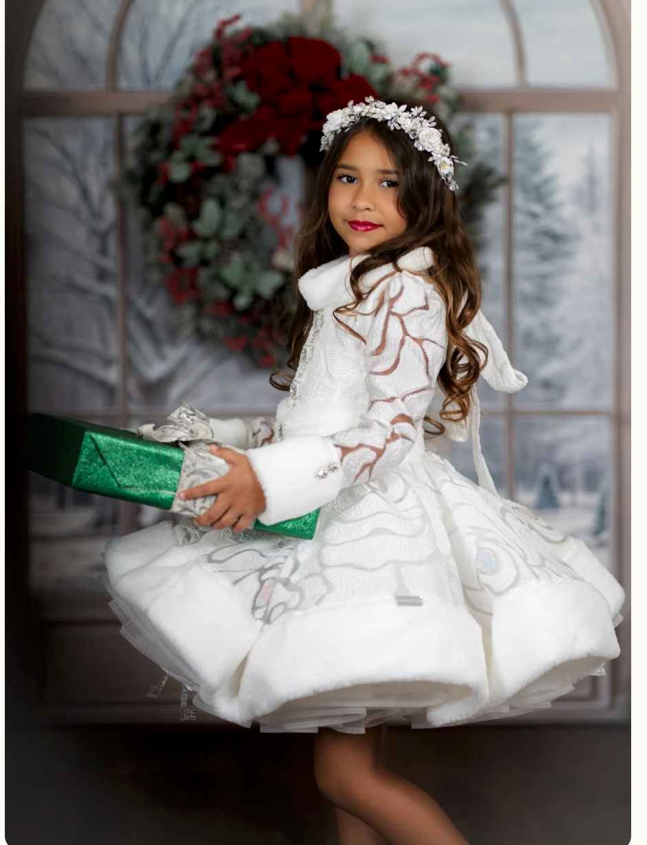 Young girl in white embroidered dress with fur trim, floral headband, holding green gift box before Christmas wreath backdrop.